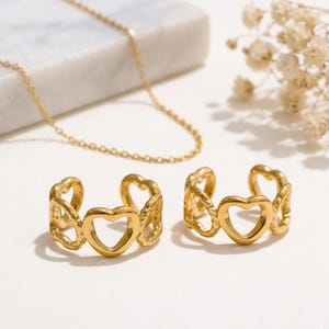 May include: Gold-colored jewelry set featuring a delicate chain necklace and two open-heart design rings. The rings have an adjustable design. The jewelry is displayed on a white surface with a marble accent and dried flowers.