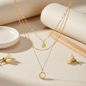 May include: A gold layered necklace with three chains. The top chain has a small sunburst pendant. The middle chain has small oval links. The bottom chain has a round, white pendant. The necklace is displayed on a neutral surface.