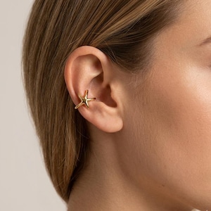 May include: A gold-coloured ear cuff in the shape of a starfish. The jewellery piece is worn on the ear cartilage. The cuff has a simple, elegant design and is made of a shiny, metallic material. The ear cuff is a unique accessory.