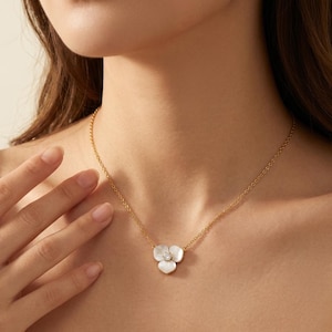 May include: A gold necklace with a three-petal flower pendant. The pendant is white with a central clear gemstone. The necklace is worn around the neck.
