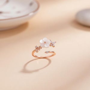 May include: A rose gold adjustable ring featuring a white flower and two butterfly accents. The ring is adorned with small, clear gemstones. The ring is open at the bottom, allowing for size adjustment. The ring is set against a soft, neutral background.