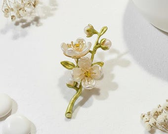 May include: A floral brooch featuring a branch with white, pearl-like flowers and buds. The petals have a pearlescent sheen, with yellow centers. The stem and leaves are gold and green. The brooch is a delicate piece of jewelry.