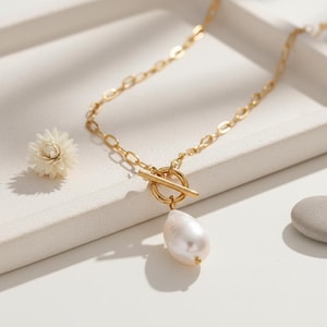 May include: A gold necklace with a large, pear-shaped pearl pendant. The necklace has a toggle clasp and an oval link chain. A small white flower and a grey stone are also visible.