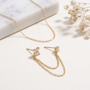 Op de afbeelding: Een gouden ketting en een paar gouden oorbellen met een ketting. Eén oorbel heeft een klein bloemmotief met witte stenen, de andere heeft een enkele heldere steen. De sieraden worden op een wit oppervlak gepresenteerd.