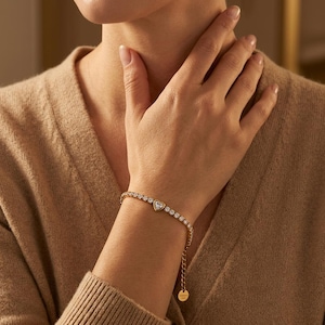 Op de afbeelding: Een goudkleurige armband met een hartvormig middelpunt en een rij ronde, heldere stenen. De armband zit om een pols, met de hand in de buurt van de nek. De achtergrond is een neutrale, warme kleur.