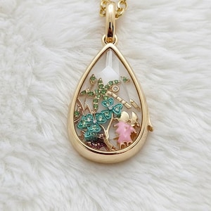 Könnte beinhalten: Goldfarbene Tropfenanhänger-Halskette mit klarem Mittelteil. Der Anhänger zeigt bunte Miniatur-Schmetterlinge, Blumen und eine Libelle. Die Halskette liegt auf einer weißen, flauschigen Oberfläche.