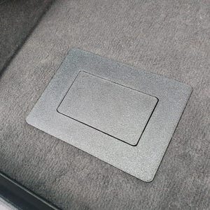 Puede incluir: Una cubierta de suelo rectangular de plástico negro con acabado texturizado, que presenta un panel central insertado. El componente está diseñado para quedar al ras de una superficie alfombrada, proporcionando un aspecto limpio y uniforme.