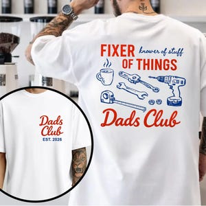 May include: White t-shirt with "Dads Club" in red script on the front and "EST. 2026" in blue. The back features the text "FIXER of THINGS" with tools and a coffee cup graphic in blue and red.