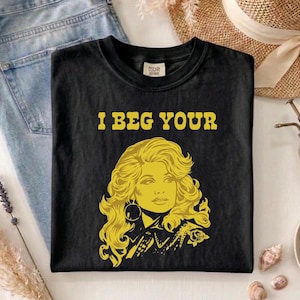 Könnte beinhalten: Ein schwarzes kurzärmeliges T-Shirt mit einem gelben Grafikdruck einer Person mit voluminösem Haar und dem gelben Schriftzug I BEG YOUR über der Illustration. Das T-Shirt hat einen Rundhalsausschnitt und eine Standardpassform.