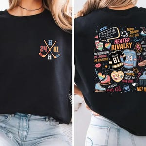 Op de afbeelding: Zwart sweatshirt met een hockeythema. De voorkant heeft gekruiste hockeysticks, de nummers 24 en 81 en de letters H en R. De achterkant heeft een kleurrijke afbeelding met tekst, waaronder "Heated Rivalry".