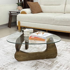 May include: A modern coffee table with a clear glass top and a unique wooden base. The base is made of dark brown wood and has a distinctive curved design. Books and decorative items are placed on the table.
