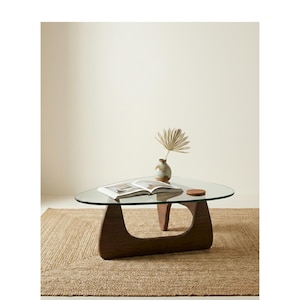 May include: A modern, triangular coffee table with a clear glass top and a dark wood base. A small vase with a dried plant and an open book rest on the table. A round coaster sits on the table.
