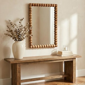 Puede incluir: Un espejo rectangular con marco de cuentas de madera cuelga sobre un banco de madera. Un jarrón blanco con ramas secas está a la izquierda del espejo. Una vela y libros están en el banco. La habitación tiene una paleta de colores neutros.