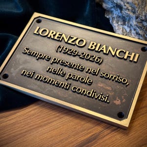 Puede incluir: Una placa conmemorativa de bronce con letras doradas. La placa dice "LORENZO BIANCHI (1929-2026)" y una inscripción italiana. La placa es rectangular con un borde dorado y está colocada sobre una superficie de madera.