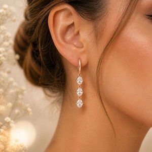 May include: Rose gold drop earrings with three oval-cut, clear gemstones. The earrings are suspended from a hoop and dangle elegantly. The image is set against a soft, blurred background. The text "GenuineGiftStore" and "Fast Shipping from USA" are visible.