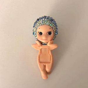 May include: A small, light-skinned figurine of a baby-like figure with a blue jeweled cap. The figurine has a smiling face with large eyes and is posed with arms raised. The body has a rectangular opening.