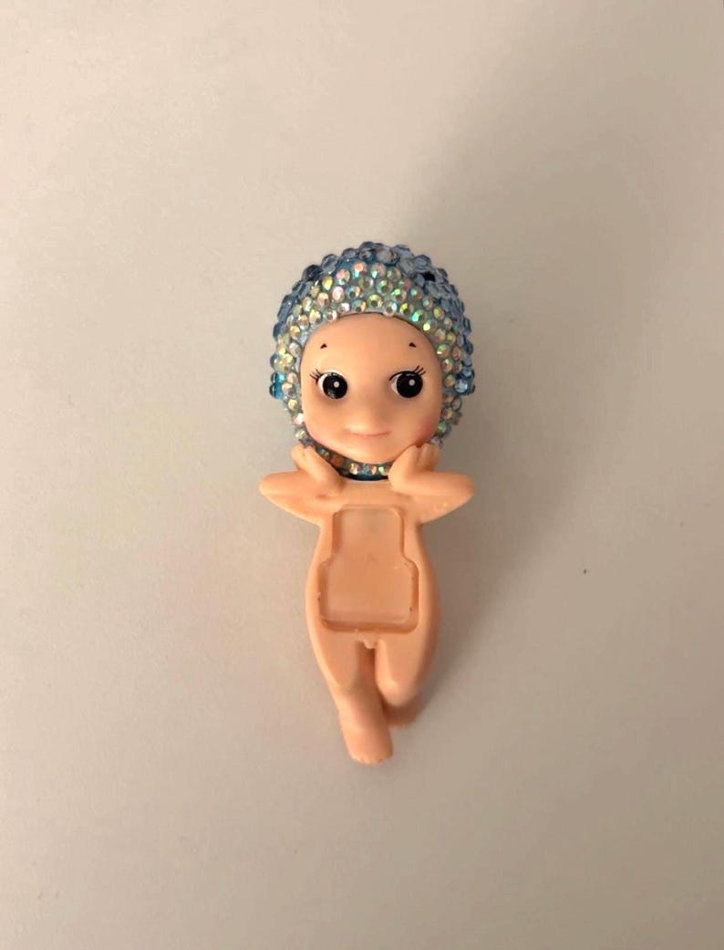 May include: A small, light-skinned figurine of a baby-like figure with a blue jeweled cap. The figurine has a smiling face with large eyes and is posed with arms raised. The body has a rectangular opening.