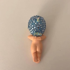 May include: A figurine of a nude human form with a blue, jewel-encrusted head. The figure is in a fetal position, with arms raised. The head is a sphere covered in light blue and iridescent gems.