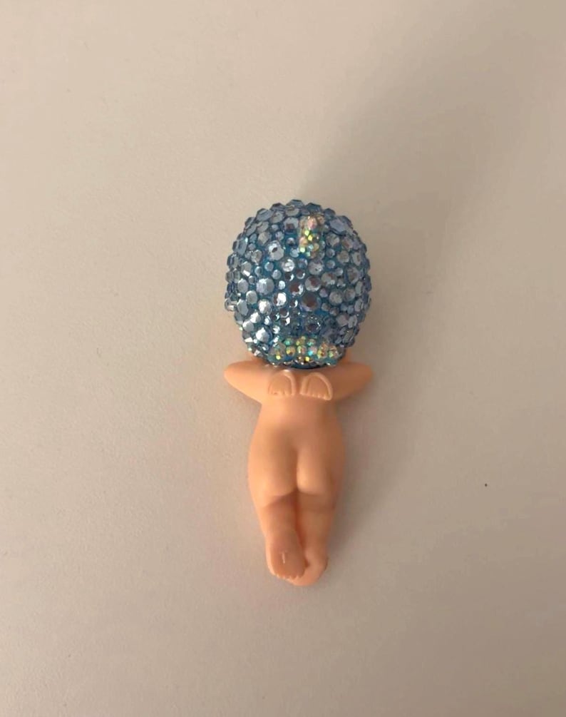 May include: A figurine of a nude human form with a blue, jewel-encrusted head. The figure is in a fetal position, with arms raised. The head is a sphere covered in light blue and iridescent gems.