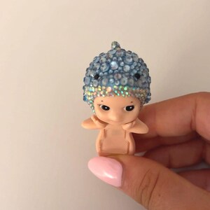 May include: A small figurine of a baby-like character with a blue jeweled hat. The figurine is held in a hand with a pink painted fingernail. The hat is covered in blue and clear rhinestones.