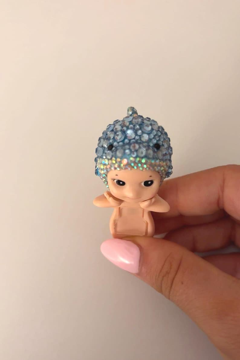 May include: A small figurine of a baby-like character with a blue jeweled hat. The figurine is held in a hand with a pink painted fingernail. The hat is covered in blue and clear rhinestones.
