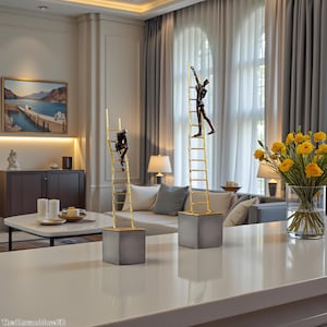 May include: Two bronze-coloured sculptures of figures climbing gold-coloured ladders. The ladders are mounted on grey concrete blocks. The sculptures are displayed on a white countertop in a well-lit room with a painting and a vase of yellow flowers.