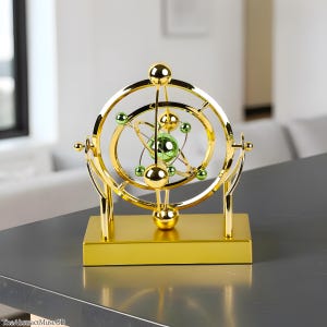 May include: A gold kinetic sculpture with a rectangular base. The sculpture features concentric rings and spheres in gold, with green spheres at the centre. The design resembles an atom model. The sculpture is displayed on a grey surface.