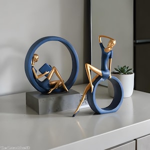 Puede incluir: Dos esculturas decorativas de mujeres en un estilo moderno. Una figura está sentada dentro de un anillo azul, leyendo un libro. La otra figura está sentada en un anillo azul. Ambas figuras tienen extremidades y cabezas doradas y visten vestidos azules.