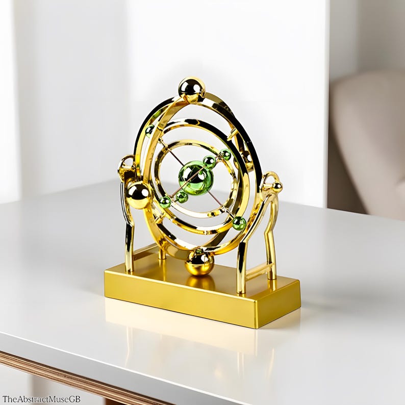 May include: A gold-colored kinetic sculpture with concentric rings and spheres. The sculpture features green and gold spheres within the rings, mounted on a rectangular base. The design is reminiscent of an orrery or a celestial model.