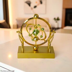 May include: A gold-colored, decorative sculpture with a spherical design. The sculpture features a central green sphere surrounded by smaller green spheres and gold rings, all mounted on a rectangular base. The AbstractMuseGB is printed on the bottom left.