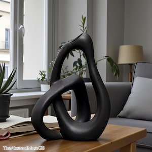 May include: A black abstract sculpture with a smooth finish. The sculpture features a looping, intertwined design, and is placed on a wooden table. The sculpture is a decorative home accent.