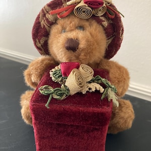 May include: A brown teddy bear wearing a burgundy plaid hat with floral accents. The bear holds a burgundy velvet box adorned with gold and red floral embellishments. The overall design is festive and decorative.