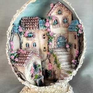 Puede incluir: Una detallada escena de pueblo en miniatura dentro de una cáscara de huevo decorativa. Las casas tienen paredes color beige, tejados marrones y están adornadas con rosas rosas. Un fondo de cielo azul completa el diseño caprichoso.