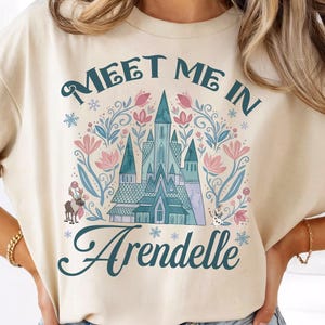 Könnte beinhalten: Beige T-Shirt mit dem Text "Meet Me In Arendelle" in türkisfarbener Schrift. Das Design zeigt ein türkisblaues Schloss, rosa Blumen und Schneeflocken. Eine skurrile Illustration einer Person auf einem Pferd ist ebenfalls enthalten.