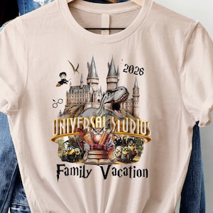 Może przedstawiać: Jasnoróżowa koszulka z grafiką przedstawiającą zamek, dinozaura i napis "Universal Studios Family Vacation 2026". Koszulka wisi na drewnianym wieszaku, z innymi ubraniami w tle.