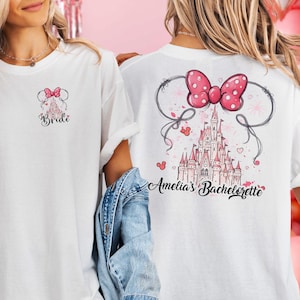 Può includere: T-shirt bianco con un design a fiocco e castello rosa. La parte anteriore dice "Bride", e la parte posteriore "Amelia's Bachelorette". Il design include un fiocco rosa a pois e un'illustrazione del castello.