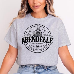 May include: A light gray t-shirt featuring a black circular design with the word "ARENDELLE" and the phrase "LET IT SNOW!" The design includes a castle, snowflakes, and the words "A PLACE WORTH MELTING FOR".