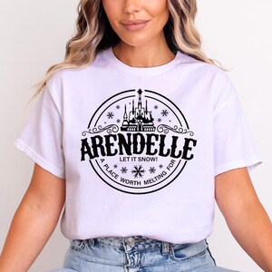 May include: White t-shirt with a black circular graphic featuring the word "ARENDELLE" above a castle illustration. The text "LET IT SNOW!" and "A PLACE WORTH MELTING FOR" are also included in the design.