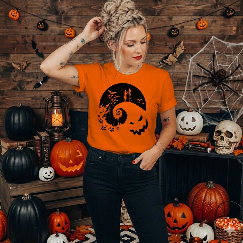 Puede incluir: Una camiseta naranja de manga corta con un estampado gr&aacute;fico negro que muestra la silueta de dos figuras sobre una colina curva, una luna grande, murci&eacute;lagos y una cara de calabaza. Se combina con unos vaqueros de color azul oscuro.