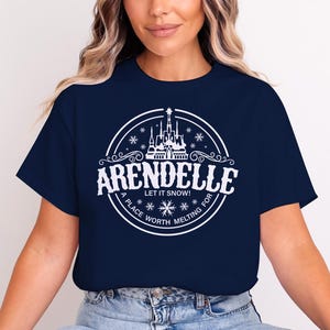 May include: Navy blue t-shirt featuring a white circular design with the word "Arendelle" and a castle illustration. The text "Let it snow!" and "A place worth melting for" are also included in the design.