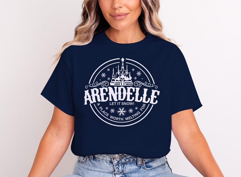 May include: Navy blue t-shirt featuring a white circular design with the word "Arendelle" and a castle illustration. The text "Let it snow!" and "A place worth melting for" are also included in the design.