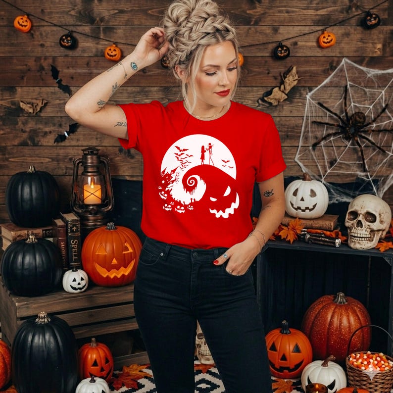 Puede incluir: Una camiseta roja de manga corta con un estampado gr&aacute;fico blanco que muestra una colina estilizada, una luna llena, murci&eacute;lagos, una casa encantada y dos figuras, con una gran cara de calabaza sonriente integrada en el dise&ntilde;o.