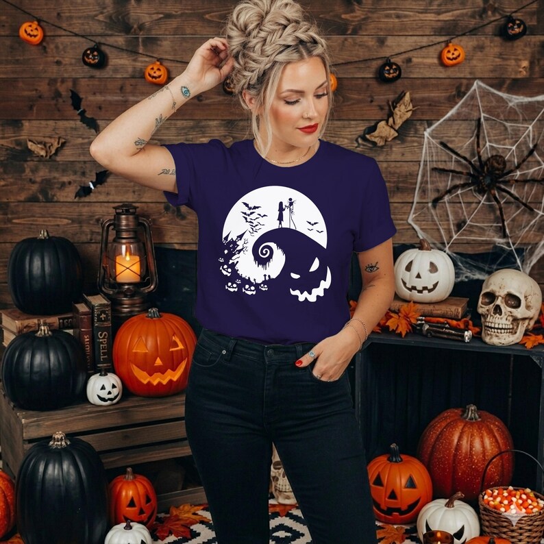 Puede incluir: Una camiseta de manga corta de color morado oscuro con un estampado gr&aacute;fico blanco de una colina en espiral, luna llena, murci&eacute;lagos, un castillo y una cara de calabaza. El dise&ntilde;o muestra la silueta de dos figuras sobre la colina.