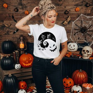Puede incluir: Una camiseta blanca de manga corta con un estampado gr&aacute;fico negro de una colina con la silueta de dos figuras, murci&eacute;lagos, un castillo y una cara grande sonriente frente a una luna llena, combinada con pantalones vaqueros azul oscuro.