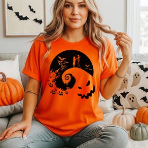 Puede incluir: Una camiseta naranja de manga corta con un estampado gr&aacute;fico negro de una colina estilizada con una espiral, un castillo, murci&eacute;lagos, calabazas y dos figuras sobre la colina bajo una gran luna circular con un rostro espeluznante.