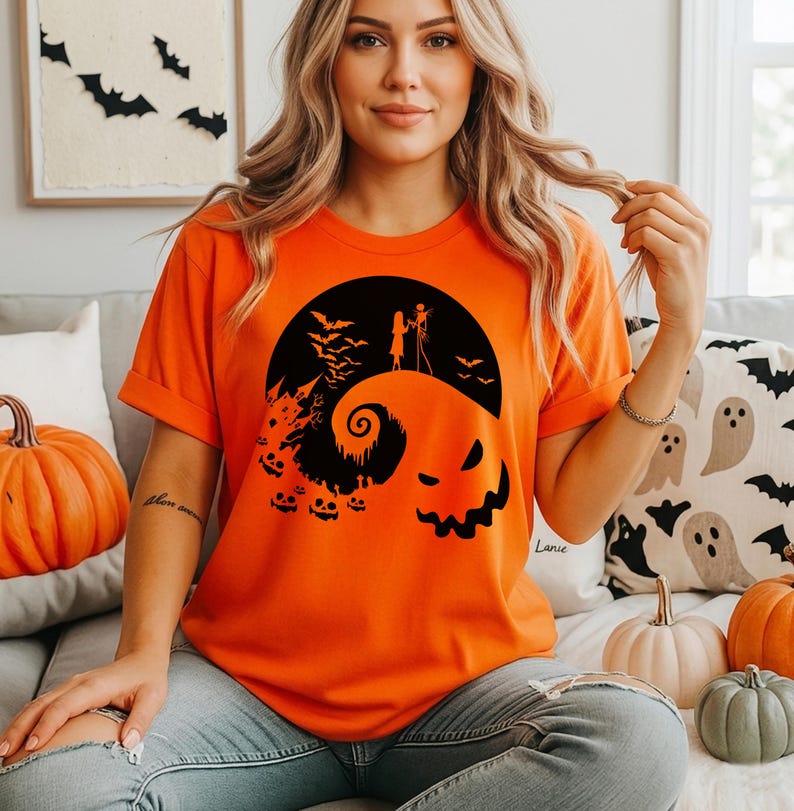 Puede incluir: Una camiseta naranja de manga corta con un estampado gr&aacute;fico negro de una colina estilizada con una espiral, un castillo, murci&eacute;lagos, calabazas y dos figuras sobre la colina bajo una gran luna circular con un rostro espeluznante.