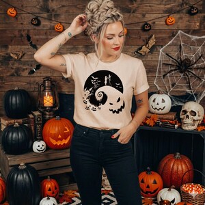 Puede incluir: Una camiseta de manga corta de color beige con un estampado gr&aacute;fico negro que muestra una colina estilizada con la silueta de dos figuras, una luna grande, murci&eacute;lagos y una cara de calabaza en la parte frontal de la prenda.