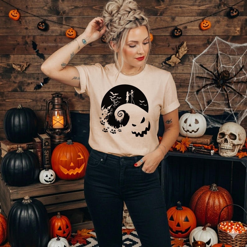 Puede incluir: Una camiseta de manga corta de color beige con un estampado gr&aacute;fico negro que muestra una colina estilizada con la silueta de dos figuras, una luna grande, murci&eacute;lagos y una cara de calabaza en la parte frontal de la prenda.