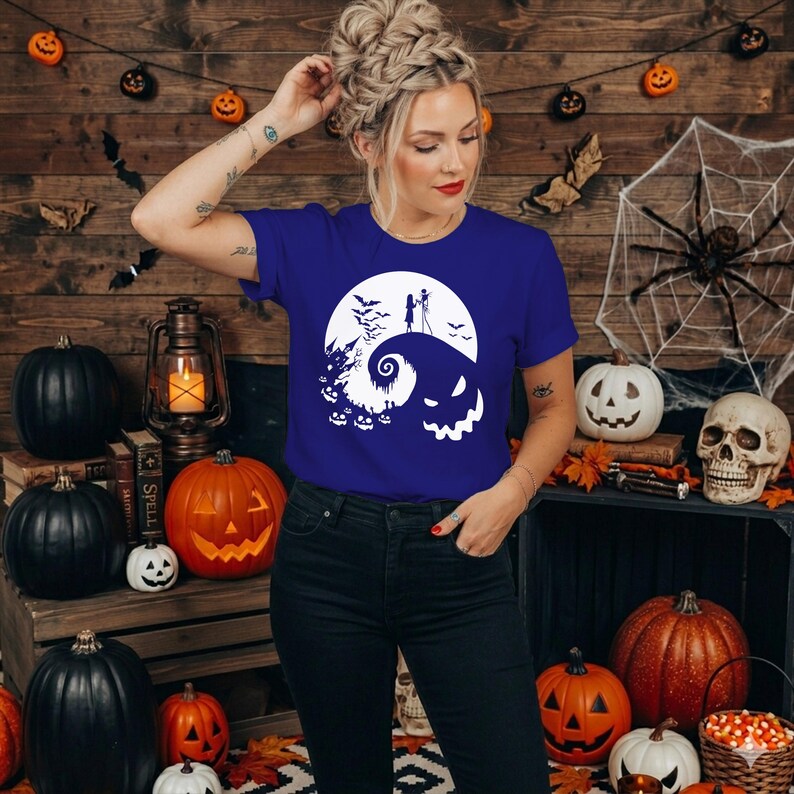Puede incluir: Una camiseta de manga corta de color azul real con un estampado gr&aacute;fico blanco que muestra una colina estilizada, una luna llena, siluetas de dos figuras, murci&eacute;lagos y una cara grande sonriente, combinada con pantalones vaqueros oscuros.