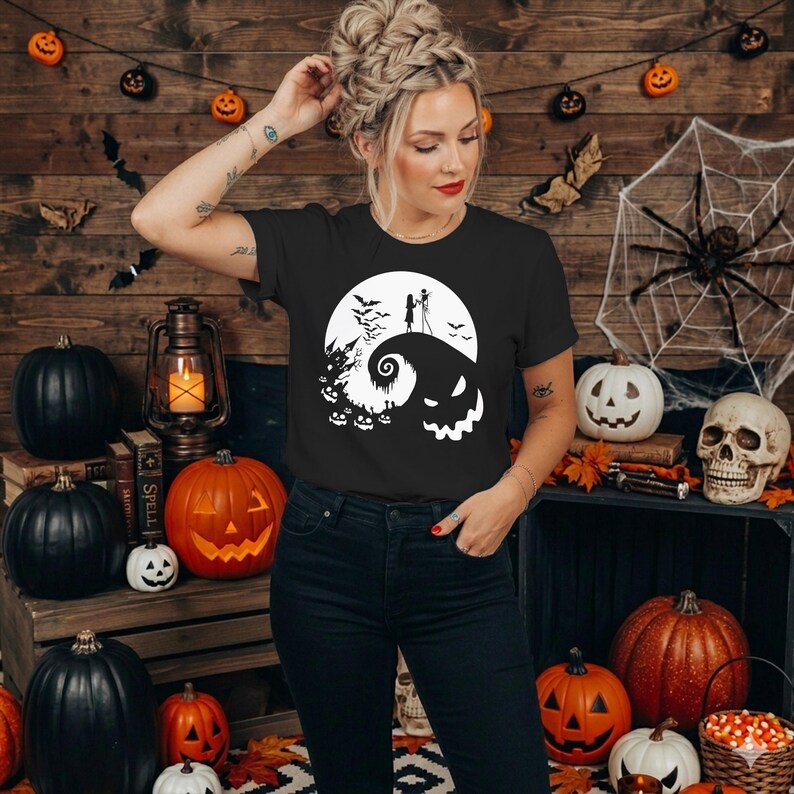 Puede incluir: Una camiseta negra de manga corta con un estampado gr&aacute;fico blanco que muestra una colina estilizada, una luna llena, murci&eacute;lagos, una casa encantada y dos figuras en la cima, combinada con pantalones vaqueros azul oscuro.